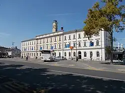 Několik autobusů na nádraží, vzadu široká budova železniční stanice