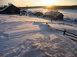 Pohled na zasněženou ves zdáli, dřevěné domy, v pozadí zamrzlá a zasněžená řeka
