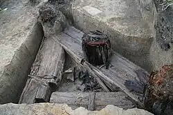 Fotografie výkopu cca půlmetru hlubokého v němž leží tři tusté dřevěné trámy tvořící pravoúhlý trojúhelník, z nejdelšího trámu trčí zbytky kůlů v něm zasazených.