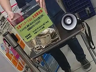 Průhledný box plný darovaných peněz v prodejně Animate, který stojí na malé stoličce a v jeho blízkosti je položen megafon