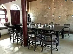 Fotografie místnosti s dřevěnou podlahou a bíle omítnutými stěnami, vlevo velké okno, vpravo na stěně velká mapa se zapíchnutými červenými a modrými praporky, uprostřed místnosti stůl, kolem něj osm židlí, na stole porcelánové hrnky a talířky, pod oknem stůl s krabicemi a skříň