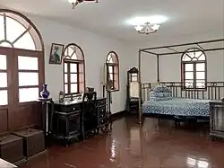 Fotografie spoře vybavené bíle omítnuté místnosti s dřevěnou podlahou, velkými okny ve stěně, psacím stolem pod okny, postelí s polštáři a truhlami vedle stolu.