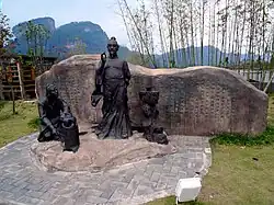 Kovové sousoší na hrubě opracované kamenné terase v parku: socha dramatika Ču Čchüana v životní velikosti, vedle něho klečící sluha s kotlíkem na kamínkách, a na kameni šálek čaje; v pozadí za kovovou plastikou; za nimi hrubá kamenná stěna s dlouhým čínským nápisem