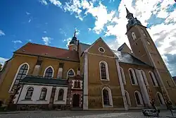 Barevná fotografie s pohledem na hlavní loď kostela, hranolovitou věž a přístavby v podobě kaplí