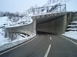 Na fotografii je silnice, která se noří do betonového, ze strany od svahu otevřeného tunelu. Nad tunelem je strmý zasněžený svah.