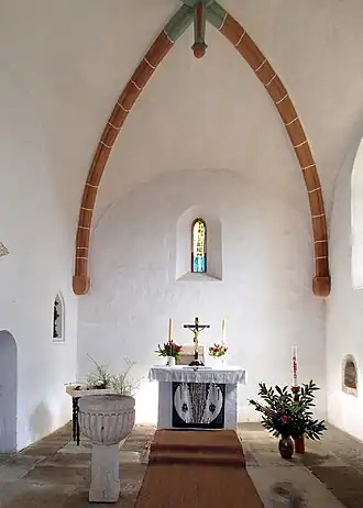 Bílé stěny, strop je klenutý, přibližně uprostřed je barevné okénko, pod ním malý oltář s ubrusy, před ním červený koberec, vlevo šedá křtitelnice, vpravo květiny