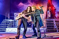 Zleva: Sascha Paeth a Tobias Sammet. Paeth hraje kytarové sólo a sešlapává kytarový pedál, Sammet stojí těsně vedle něj a pozoruje ho.