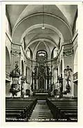 Pohled do presbytáře na černobílé pohlednici, uprostřed hlavní oltář, po stranách vítězného oblouku kazatelna a kříž s Bolestnou Matkou Boží, v popředí lavice