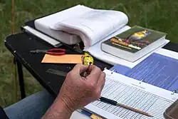 Na fotografii lze vidět ruku třímající malého ptáka, na pozadí je stůl s ornitologickými publikacemi a pomůckami