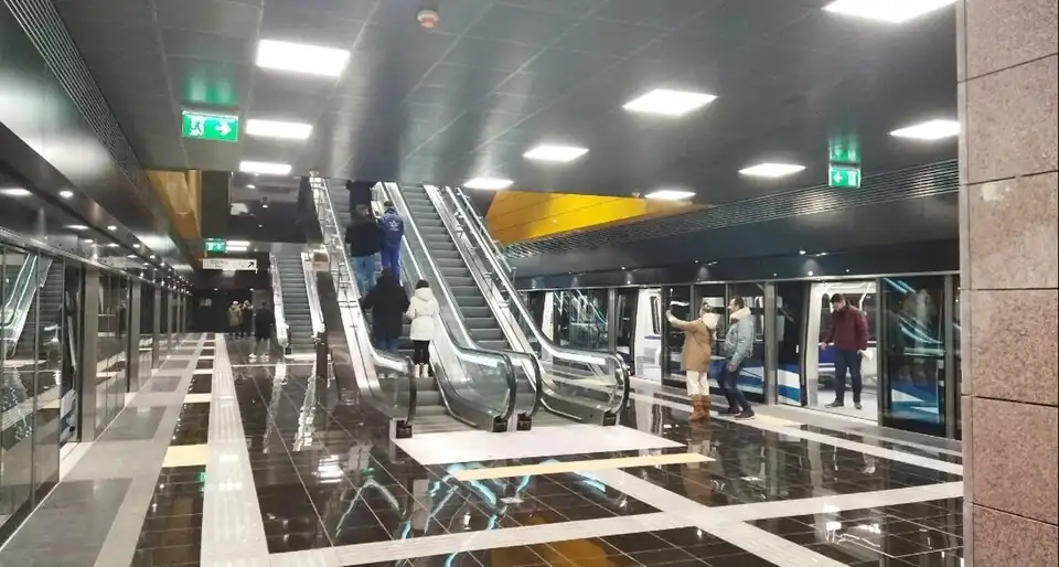 Pohled na nástupiště podzemní stanice metra. Po obou stranách jsou prosklené posuvné dveře nástupiště, vpravo jsou otevřené a lidé vystupují z vlaku. Interiér působí moderně, uprostřed nástupiště jsou vidět dvě dvojice eskalátorů, které vedou nahoru k povrchu. Podlaha z lesklých čtvercových dlaždic černé a bílé barvy. Šedý nižší strop z panelů, do něj jsou zakomponovaná světla.