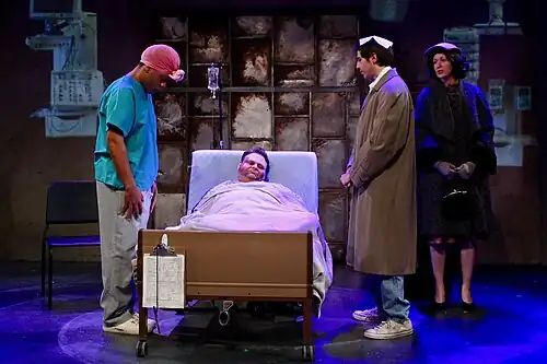 Na nemocniční posteli leží zesnulý Roy Cohn, kolem něho stojí Belize, židovský chvalozpěv.Louis a duch Ethel Rosenbergové, kteří recitují kadiš,