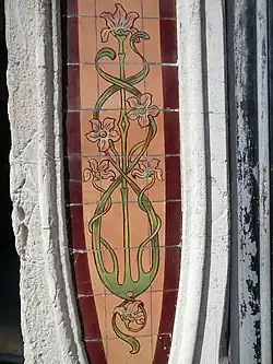 Secesní keramický obkladový panel z kachlů s rostlinným ornamentem. Na červenohnědém podkladu je vyobrazen stylizovaný rostlinný motiv se zelenými stonky a listy, které vytvářejí plynulé, vlnité křivky a uzavřené tvary. Na stoncích jsou růžové nebo červené květy, ve spodní části je malý rostlinný výhonek. Panel je vertikálně orientovaný s oválným tvarem a je zasazen mezi světlými architektonickými prvky.