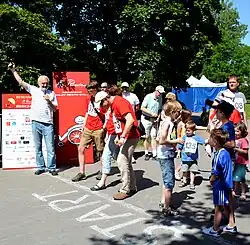 Asfaltová cesta, na níž je patrná bílá čára a nápis „Start“. Za čárou se k běhu připravují sportovci a v pozadí stojí Zdeněk Svěrák třímající v levé ruce mikrofon, do něhož hovoří, a v pravé ruce míří do vzduchu startovní pistolí.