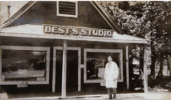 Na černobílé fotografii je muž oblečený jako malíř, stojící před dřevěným domem, na kterém je cedule s nápisem BEST´S STUDIO.