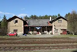 Patrová staniční budova s kolejištěm před ní. Nalevo před budovou parkuje červený osobní automobil Škoda Felicia.