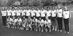 Aranžovaná skupinová sportovní fotografie fotbalového mužstva 1. FC Lok Leipzig z roku 1976, stejný postoj všech hráčů se založenýma rukama za zády a uniformované pózy sedících vpředu. Fotograf nestál v ose fotografované skupiny, což má za následek zkreslení velikosti hráčů a mírně šikmou kompozici.