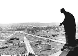 Černobílá fotografie zachycující barokní sochu na ochozu kostela, za níž jsou vidět prázdná prostranství porostlá trávou