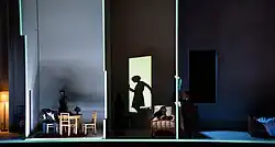 Záběr z operetního představení na šířku. Zobrazuje lidé ve třech místnostech. V první gestikuluje muž sedící na židli rukama, pozoruje ho žena. V druhé místnosti jsou tři postavy, muž sleduje ženu ležící na posteli a z druhé strany do dveří vstupuje další žena. Ve třetí místnosti s postelí se o stěnu opírá muž.