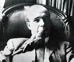 Černobílá fotografie představující hlavu a ramena muže v obleku s kravatou, na jehož hrudi sedí živý papoušek karolinský