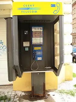 Na velkoměstské ulici stojící trojstěnný přístřešek s telefonním automatem. Přístřešek je nahoře označený žlutou cedulí s logem tvořeným modrými velkými písmeny zapsanými slovy ČESKÝ TELECOM (druhé slovo pod prvním), oddělenými třemi čárami přecházejícími zleva doprava z modrých v zelené.