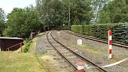 Dvě souběžně vedené úzkorozchodné železniční koleje. Vlevo od tratě lemované ostříhaným živým plotem z jehličnatých stromů. Mezi kolejemi a kolem nich železniční značky.