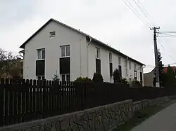 Bíle omítnutá patrová budova se zahradou rozkládající se kolem objektu ohraničená dřevěným plaňkovým plotem.