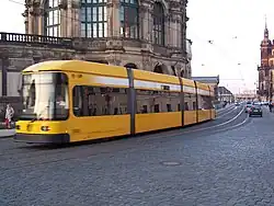 Barevná fotografie s pohledem na projíždějící žlutou tramvaj, za níž je jeden z pavilónů Zwingeru