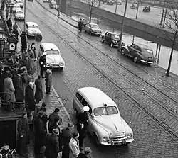 Černobílá fotografie ulice s nábřežím vodního kanálu, po ulici jede kolona sanitek.
