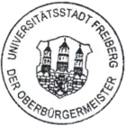 Černobílá fotografie s kulatým razítkem, lemovaným nápisem, uprostřed je městský znak