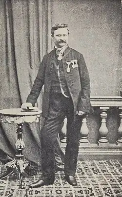 Černobílá fotografie zachycující muže s krátkými vlasy a knírem, který se opírá o malý stůl.