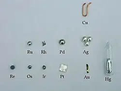 plíšky, zrnka a drátky vyrobené z jednotlivých ušlechtilých kovů srovnané ve tvaru periodické tabulky prvků. Zleva shora měděný zahnutý drátek, zrnko ruthenia, odlitek rhodia, zrno palladia, tři zrnka stříbra, zrnka rhenia, osmia a iridia, plíšek platiny, pásek zlata a ampulka rtuti