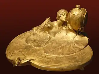 Zlatý secesní kalamář zobrazující nahou ženskou bustu na hladině s listy leknínu na kruhovém podstavci. Postava drží nádobku na inkoust. Předmět je vyfotografován na tmavě červeném pozadí.
