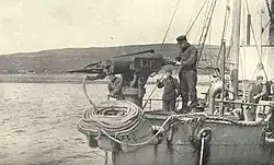 Stará černobílá fotografie přídi lodi, kde je instalováno mohutné, asi metr a půl vysoké zařízení - dělo - s nabitou harpunou. ze které vede silné lano, které je svinuto pod dělem. U děla (jeho rukojeti) stojí muž, který může dělo ovládat na otočném podstavci.