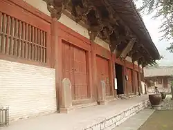 Fotografie stěny budovy, na nízké kamenné terase, ubíhající zleva doprostřed, bližší část z cihel, většina však dřevěná