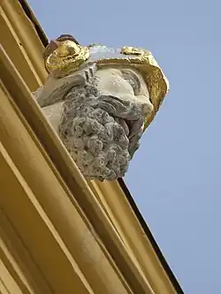 Barevná fotografie s pohledem zespodu na šikmo ubíhající římsu, na níž je plastika zobrazující mužskou hlavu s vousy