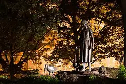 Pohled na kašnu ve večerních hodinách. Kašna i stromy jsou zahalené stínem, v pozadí žluté světlo z lamp.