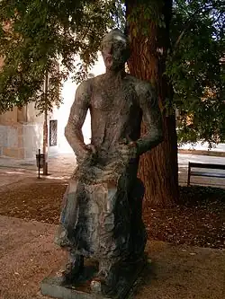 Bronzová socha sedícího muže s rukama v klíně. Za sochou se nachází jehličnatý strom, za ním pak vlevo lze vidět část domu.