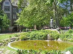 Fotografie kašny, socha je natočena lehce levým bokem. Je vidět celé jezírko, vzadu nad ním je umístěna socha dívky se třemi husami.