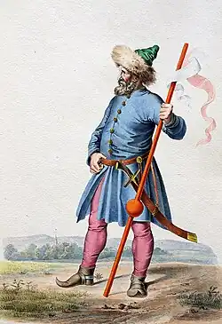 Starší muž s plnovousem oblečený v modrém kabátě s kalpakou na hlavě, červenorůžovými kalhoty a hnědé boty s úzkou špičkou. V ruce drží prapor a dívá se směrem doleva.