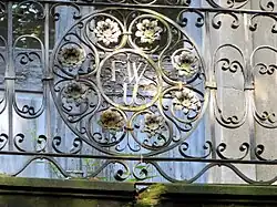 detail kování mříže balkonu, uprostřed se dvěma kruhy. V mezikruží osmero květů, ve vnitřním kruhu zdobené iniciály FWU