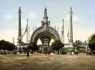 Monumentální vstupní brána Světové výstavy v Paříži z roku 1900 s výrazným obloukem a dvojicí vysokých dekorativních věží. Před bránou jsou vidět lidé, koňské povozy a vlajky. V pozadí je vidět část Eiffelovy věže.