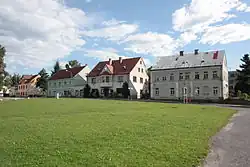 Pohled přes travnatou plochu na patrové domy stojící v řadě a mající mezi sebou proluky. Před domy vede vozovka a nad nimi je modré nebe s bílými mraky.