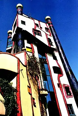 Budova s charakteristickou organickou architekturou Friedensreicha Hundertwassera. Bílá fasáda je zdobena červenými nepravidelnými křivkami, doplněná modrými sloupy se zlatými kopulemi na vrcholu. Budova má asymetrické balkony s vegetací a typické Hundertwasserovy prvky jako nerovné linie, pestré barvy a odmítání pravých úhlů. Snímek je pořízen z podhledu proti jasnému modrému nebi.