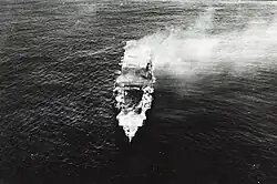 Černobílá letecká fotografie letadlové lodi, pohled zepředu. V přídi zeje velký otvor, zaujímající první třetinu lodi.