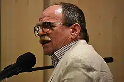 Muž v brýlích hrající na klávesový nástroj a zpívající do černého mikrofonu