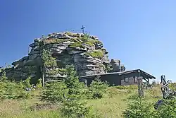 Pohled na vrcholovou skálu na hoře Jizera. Na skále stojí kříž a zábradlím obehnaná vyhlídka. Před skálou pak dřevěná bouda.