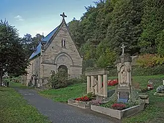 Vlevo hřbitovní kaple, stavba s trojúhelníkovým štítem z šedohnědého kamene, v popředí zdobené náhrobky se sochami a květinami, v pozadí stromy a nad nimi modrá obloha