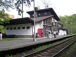 Železniční zastávka v obci Křivoklát na trati Rakovník – Beroun. Asymetrická budova, vystavěná pomocí kombinace zdiva a dřevěných prvků, s listnatým lesem v pozadí. V popředí obrázku se nachází moderní nástupiště z betonových panelů a traťová kolej.