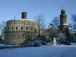 Barevná fotografie s pohledem na masivní gotickou obrannou věž a širokou válcovou baštu někdejšího opevnění
