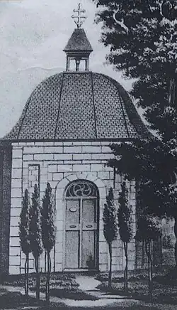 Černobílá kresba, pohled na jednu ze tří stran kaple, ze střechy kryté břidlicí vybíhá lucerna zakončená křížem, ve zdi jsou vsazeny půlkruhově zakončené dveře, vpravo od kaple větve vzrostlého stromu, před kaplí je přístupová cesta lemovaná šesti mladými stromy